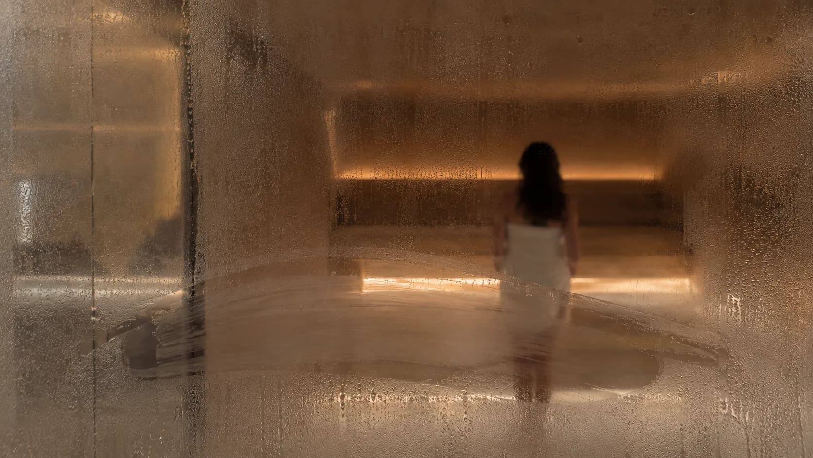 A woman in a towel behind steamed glass, a hand-wiped arc clearing the fog